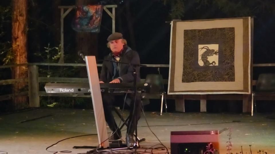 Viktor at a keyboard on an outdoor stage beside a framed artwork, playing Imagine Peace on Earth.