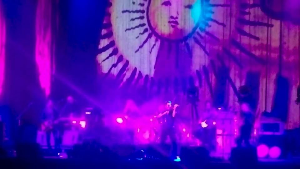 A stage lit in purple and pink with a large sun banner behind the band at the Greek Theatre.